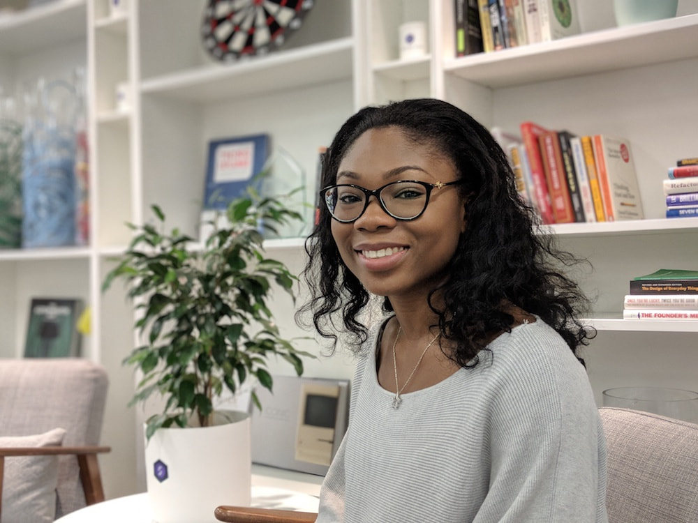 Mariam Ogunbambi smiling wearing a grey top sitting in front of a bookcase.