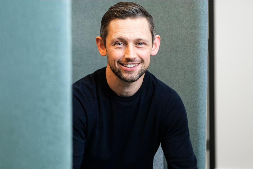 Man smiling directly to camera, wearing a black jumper. He has grey hair and is indoors.