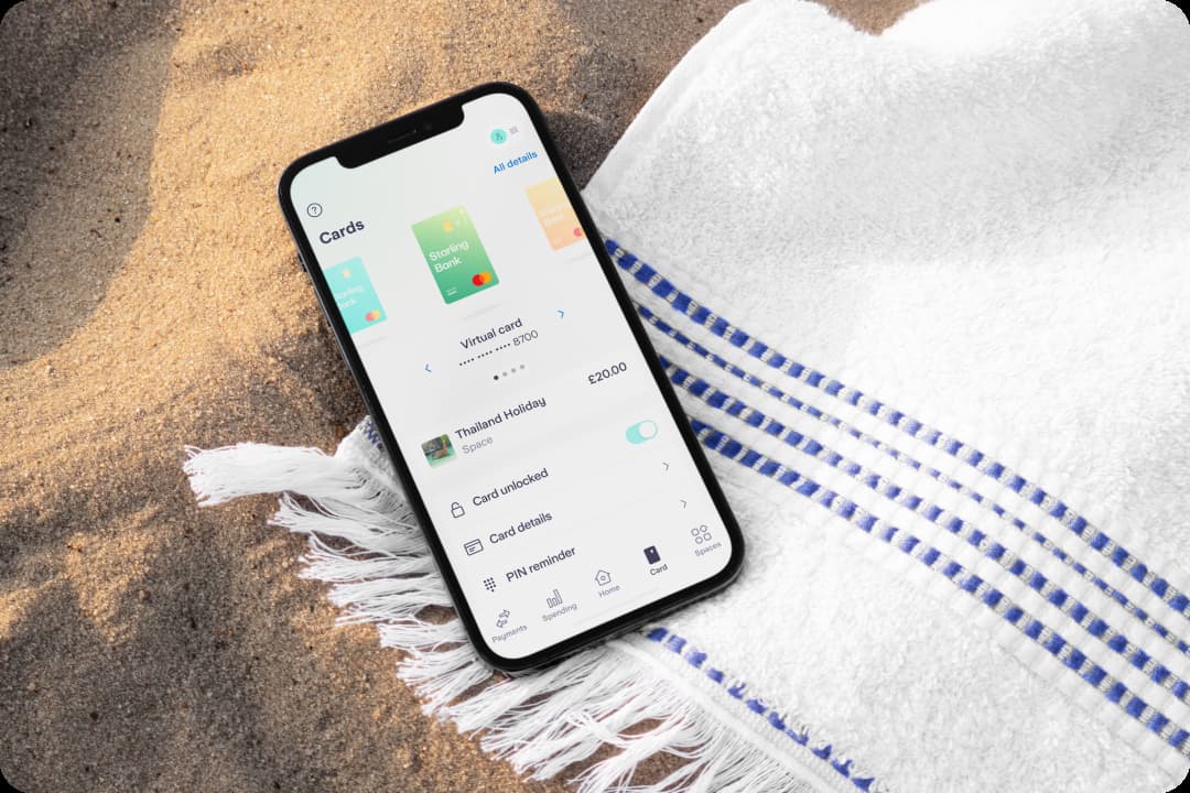 An iPhone shows the Starling app, open on the virtual card screen. The phone is on a sandy beach resting on a white and blue fringed towel.