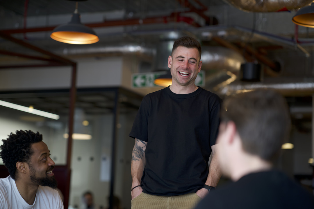 Dan Hogan, a white man in his 30s wearing a black tee-shirt laughs with colleagues. In the left hand corner, a black man in his 30s looks at Dan. The third colleague has his back to the camera.