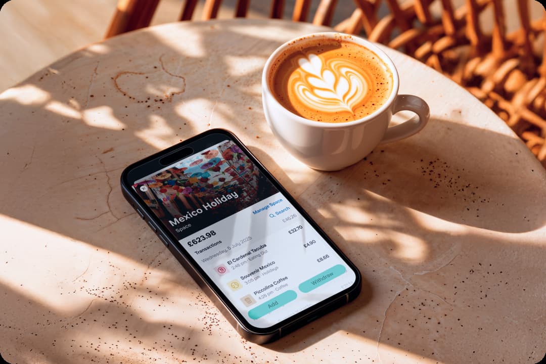 A phone showing the Spaces section of the app rests on a table next to a coffee. The Space is called Mexico Holiday and shows various transactions made from the Space.
