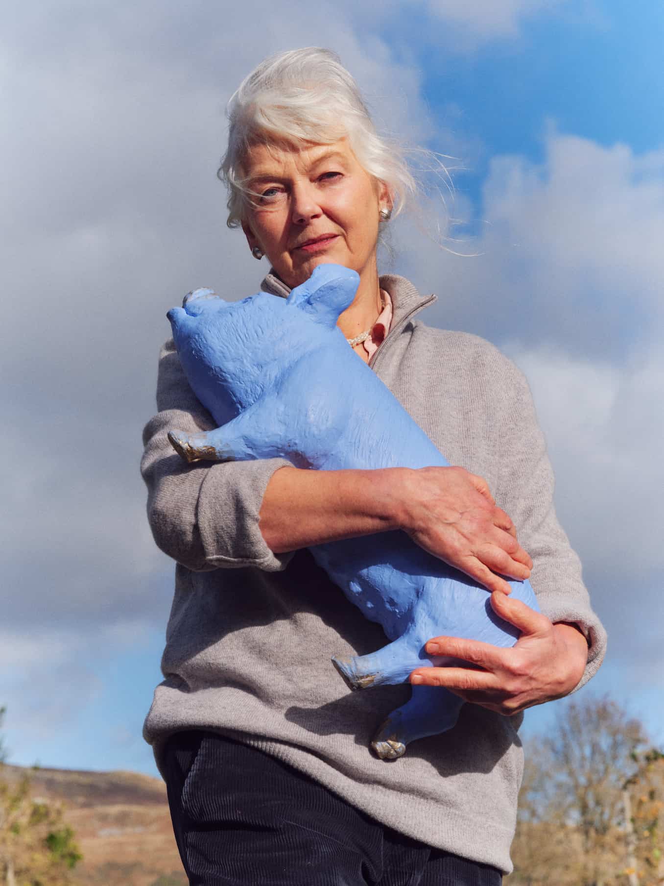 Sculptor Deirdre Nicholls holding one of her sculptures outside. The sculpture is a sky blue pig, which isn't quite lifesize.
