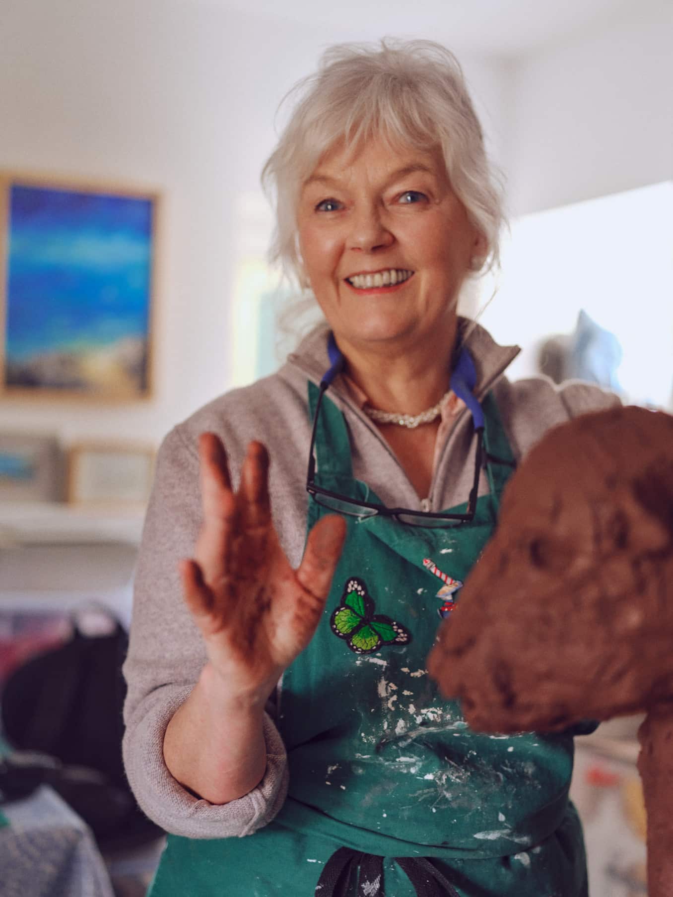 Sculptor Deirdre Nicholls looking to camera in her studio. She has clay on her hands and the sculpture she is working on is in the foreground.