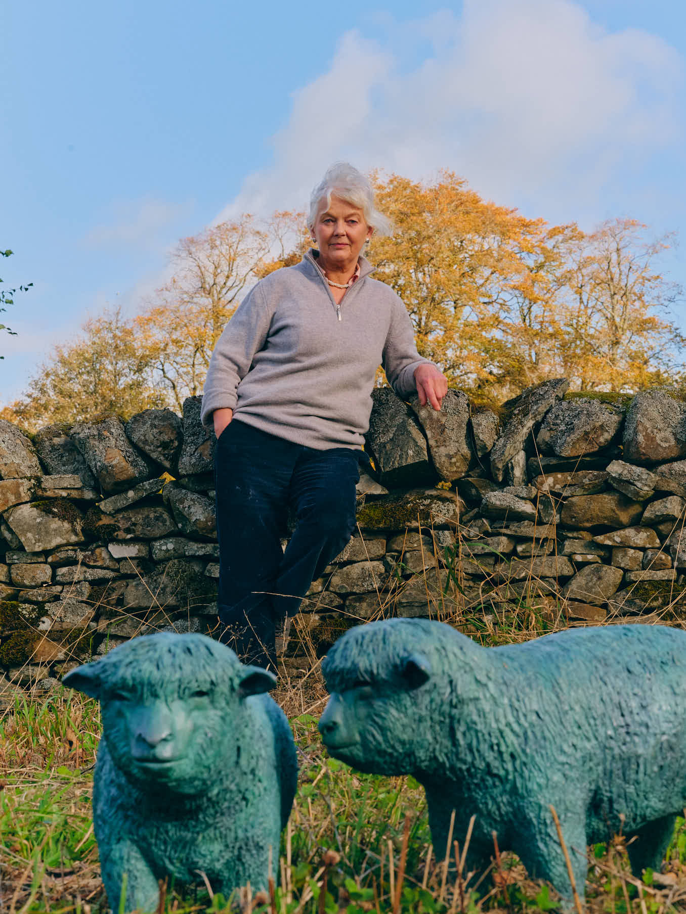 Sculptor Deirdre Nicholls standing by a stone wall, behind two of her bronze sheep sculptures