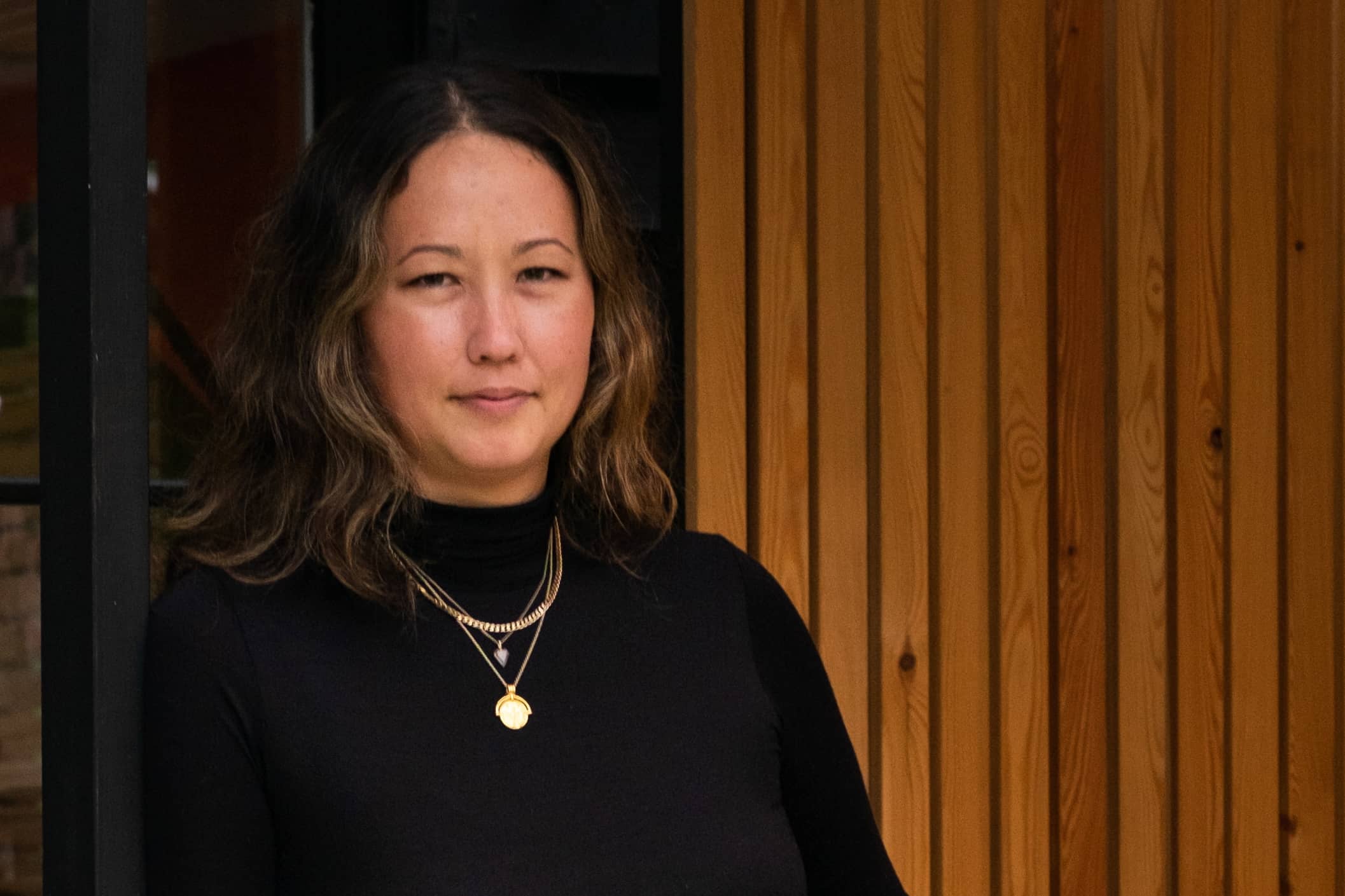 Woman looks to camera near some outdoor wood panelling. She is wearing a black polo neck and gold jewellery.