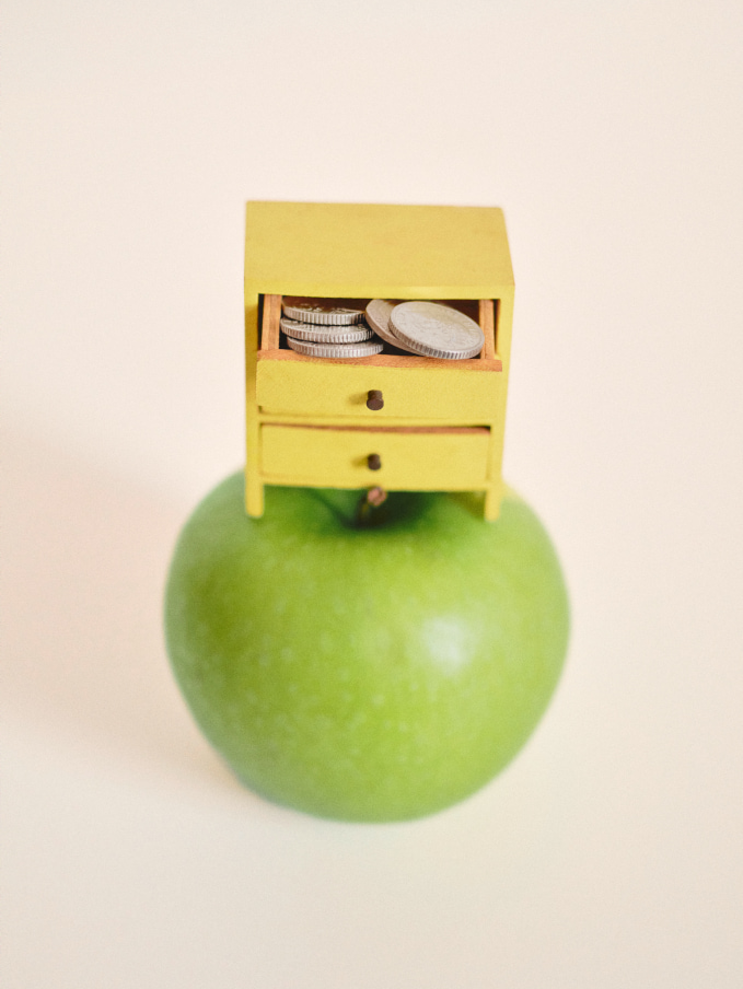 Creative photograph of a doll's house sized chest of drawers filled with coins sitting on top of a green apple kitchen set surrounded by chicken eggs